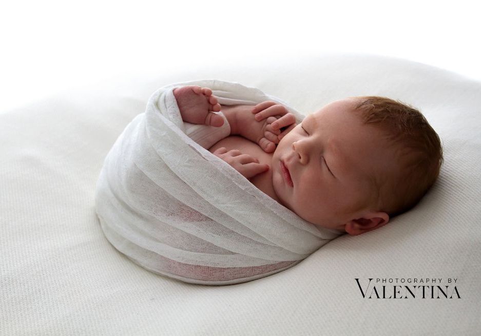 Newborn baby curled up on his back holding his little foot. He's laying on a white blanket and had a white wrap around. Photo taken during a newborn shoot in London.