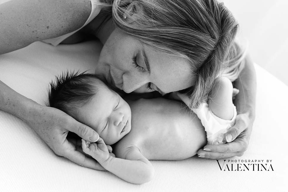 A mother lying beside her newborn baby, gently holding the baby's hand and resting her face close to the baby's head.