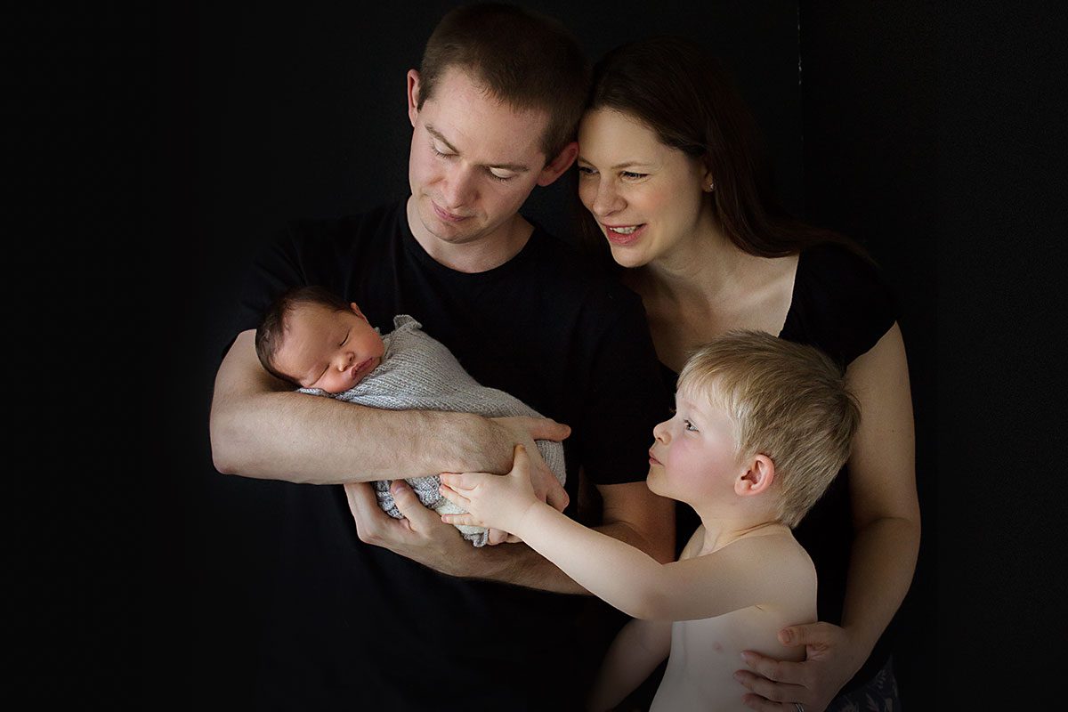 portrait of family of 4 on a black background. dad is holding newborn and little brother and mum are looking at him