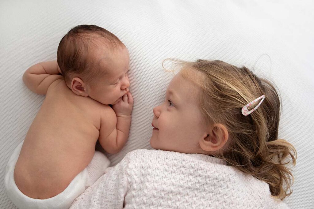 portrait of newborn and big sister laying on a white blanket and looking at each other