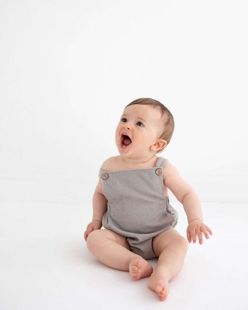 Laughing baby in a grey romper sitting against a white background – minimal studio portrait by Photography by Valentina