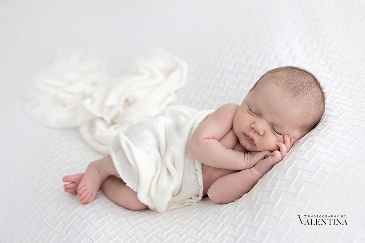 baby picture: newborn is laying on his side and sleeping on a white blanket with a soft white warp to keep him warm.