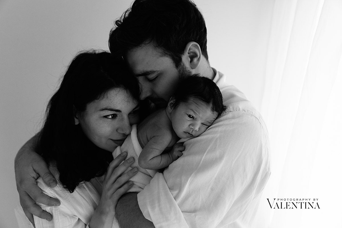 black and white family portrait. dad is holding newborn baby on his shoulder and mum is leaning into both.