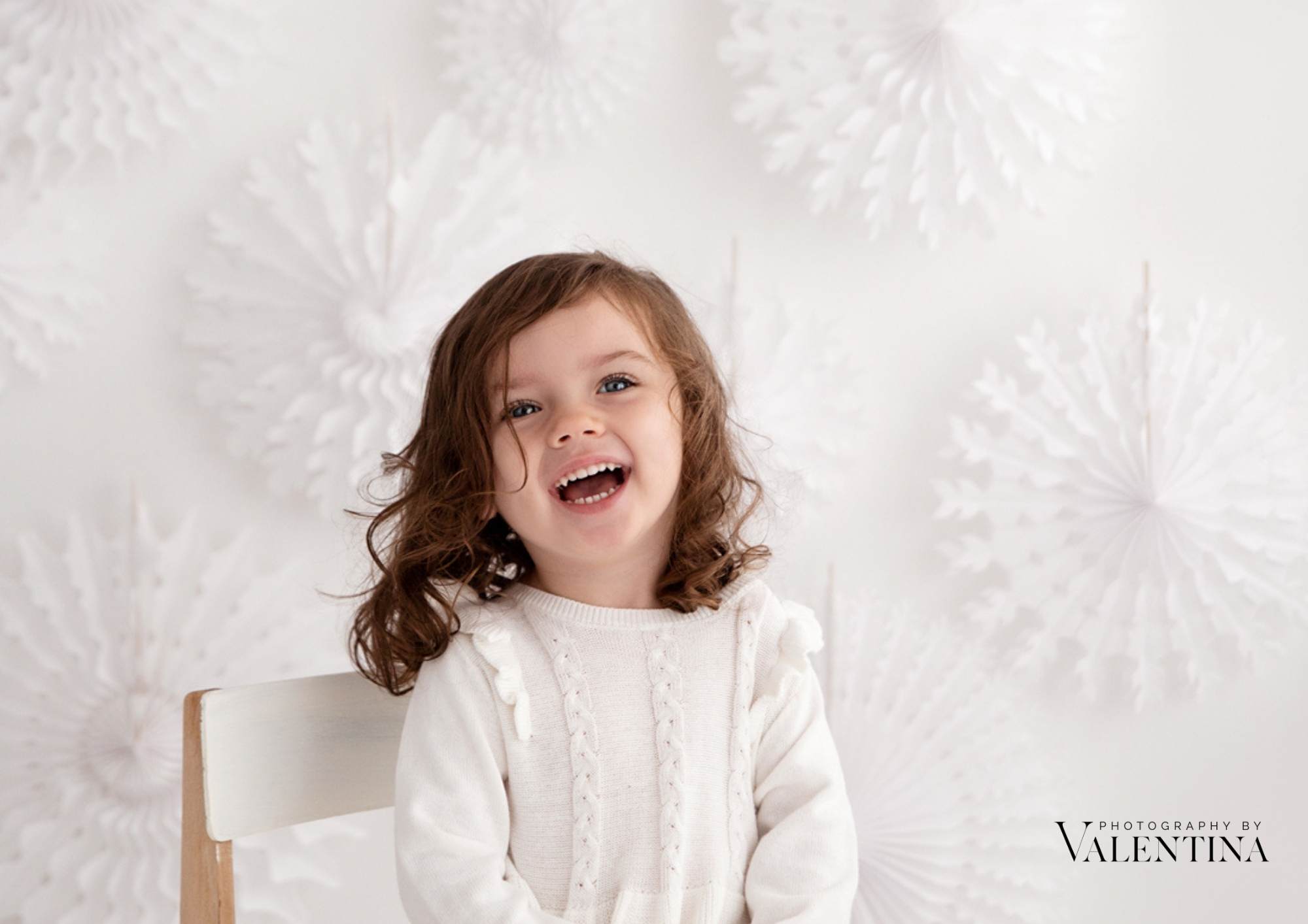 Smiling young girl with curly hair sitting on a white chair, wearing a cream knit jumper, in front of a festive white snowflake backdrop during a holiday-themed photoshoot