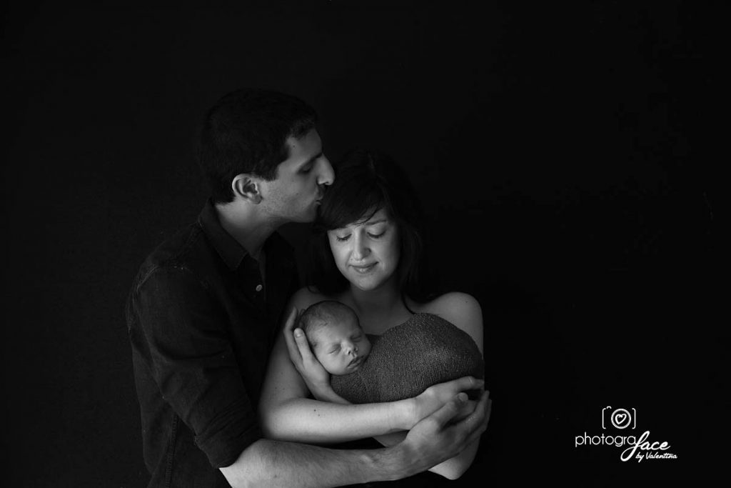 Black and white image of a mother holding her newborn while the father kisses her forehead.