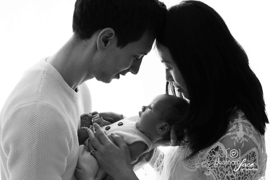 Black and white photo of new parents tenderly gazing at their newborn baby in a softly lit embrace.