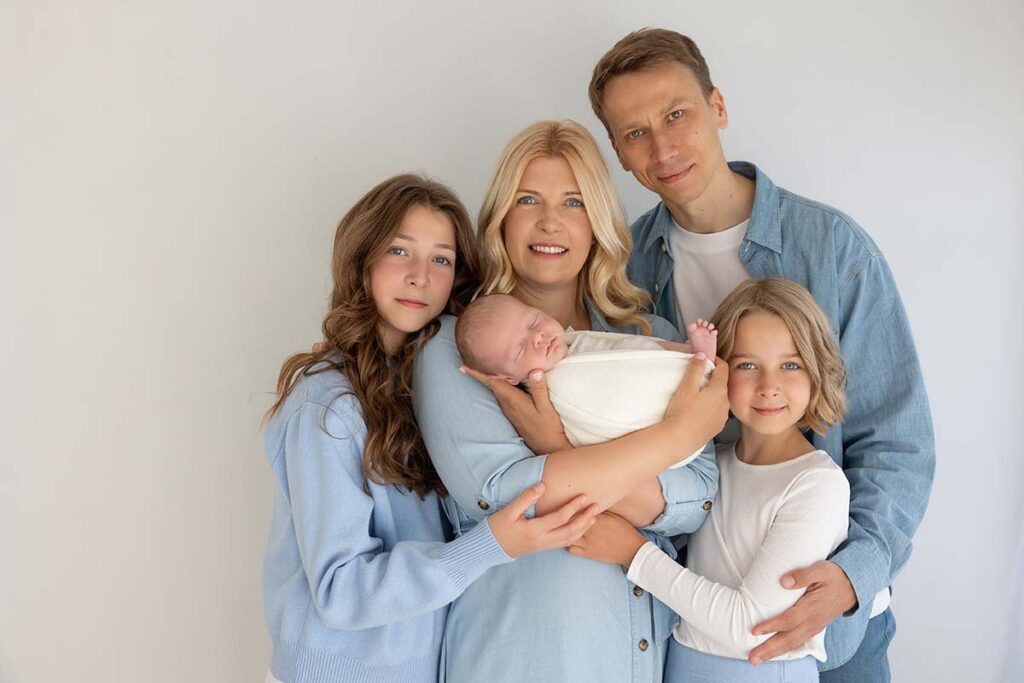 Smiling family holding their newborn baby during a photoshoot in a London studio. Parents and siblings dressed in soft blue and white outfits, showing a calm and loving connection.