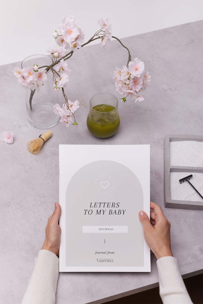 Hands holding the Letters to My Baby journal on a soft-toned table with cherry blossoms and a glass of matcha, evoking a calm spring mood—perfect for mums expecting a March baby.