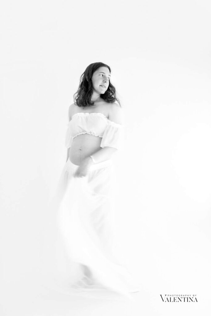 Elegant black and white maternity photography showing pregnant woman in flowing white dress during professional photo shoot for pregnant woman