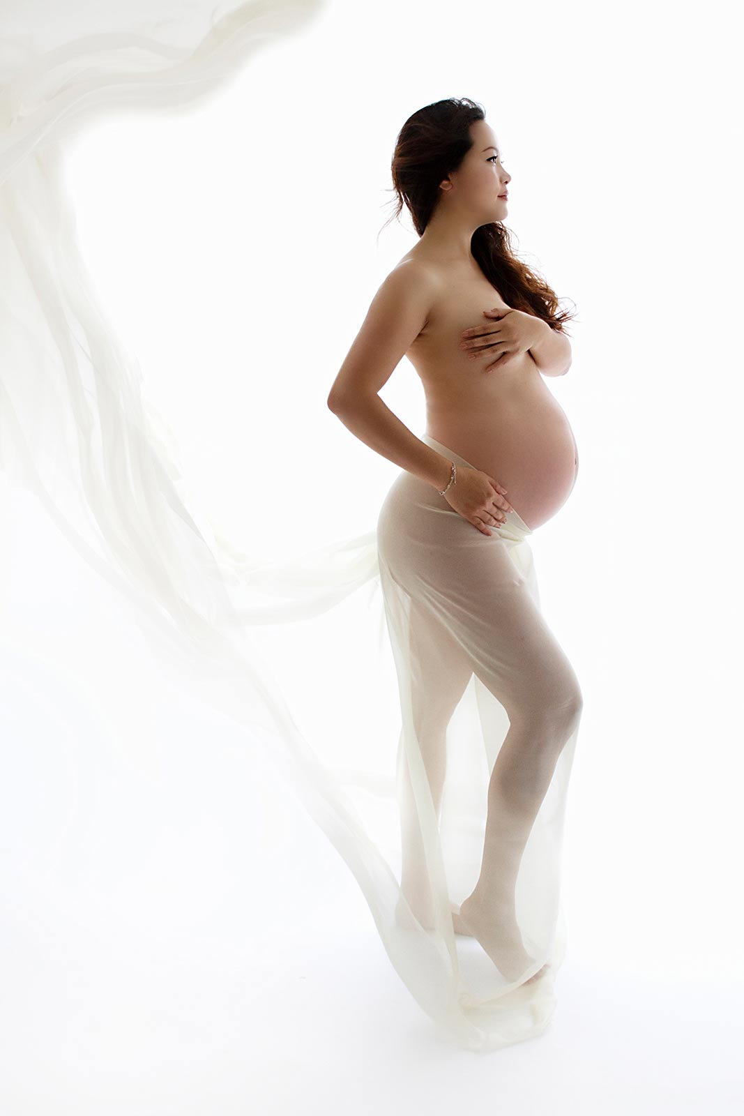 Pregnant woman standing in profile with flowing fabric, nude maternity portrait on white background, ethereal and serene