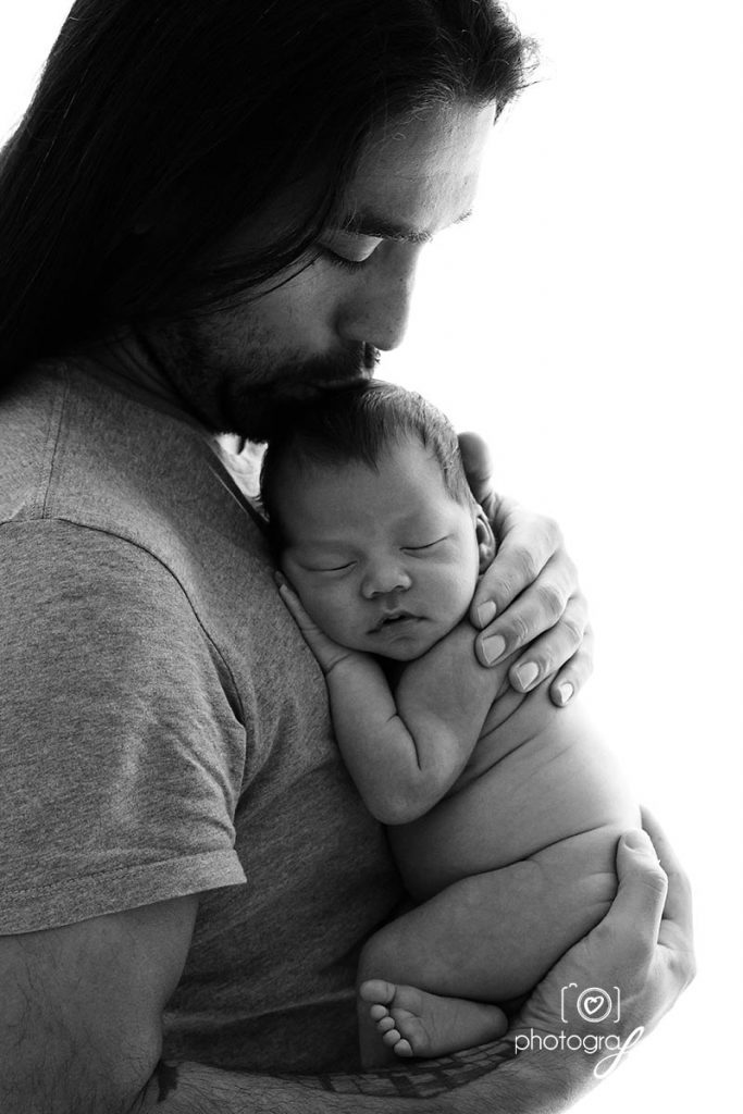 Black and white photo of a bearded dad holding his newborn close to his chest with eyes closed.