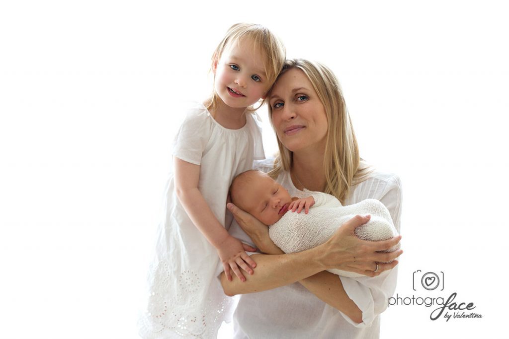 A mother holding her newborn while the older sibling stands beside her smiling, all in soft white tones.