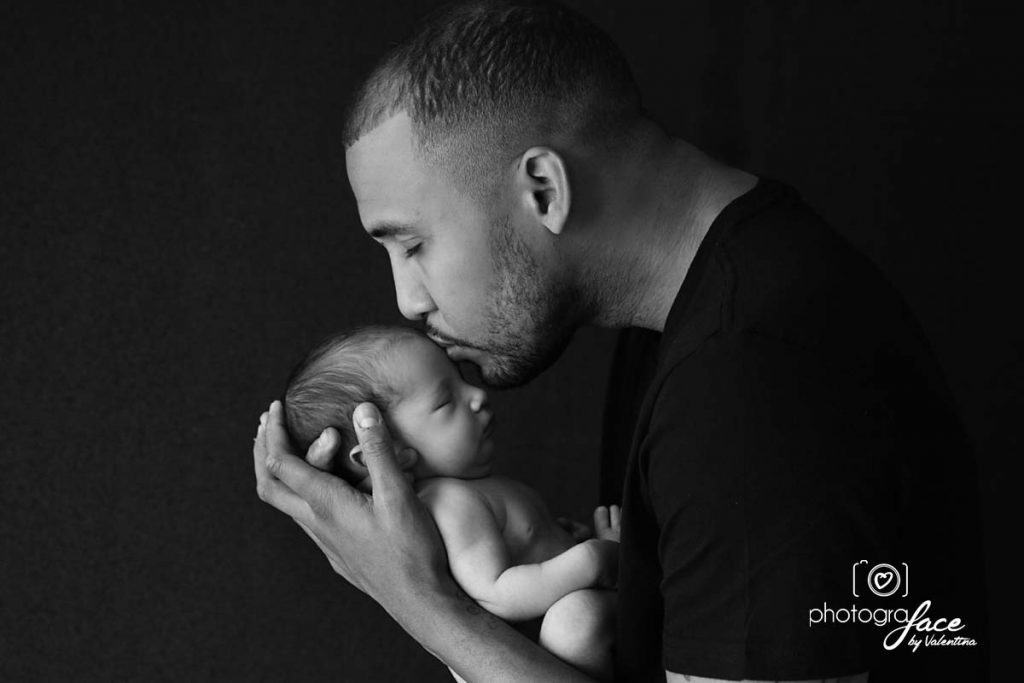Black and white photo of a father gently kissing his newborn’s head while cradling the baby in his hands.