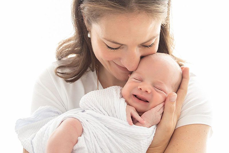 Newborn photographer in Richmond, London – mother cuddling and smiling at her newborn baby wrapped in a white blanket, captured in natural light.