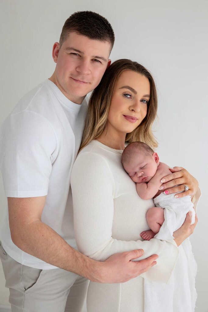 Parents holding their newborn baby in a light-filled London studio. The family is dressed in simple white tones, creating a timeless and peaceful portrait full of warmth and connection - what to wear for your newborn shoot