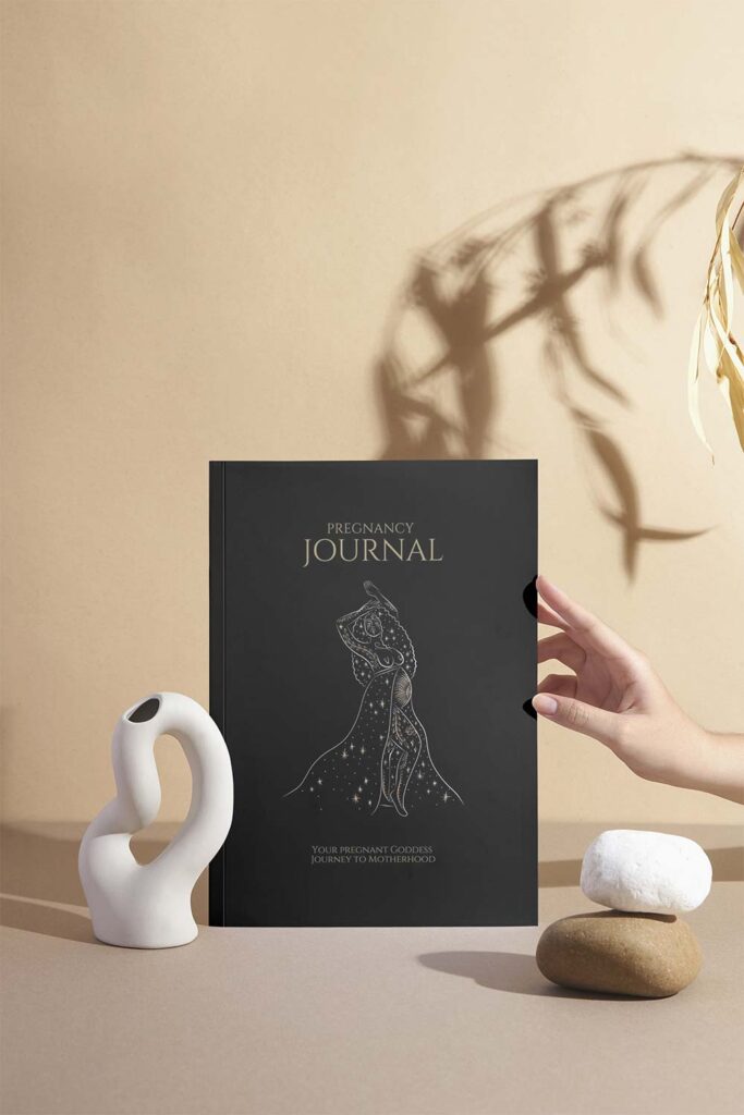 A hand gently holding a black pregnancy journal titled "Your Pregnant Goddess Journey to Motherhood" with a minimalist white sculpture and stacked stones beside it, all set against a neutral beige background with soft botanical shadows.