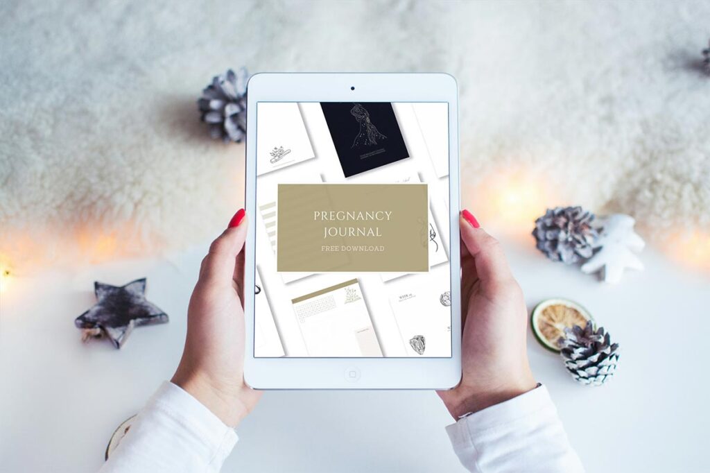 Hands holding a tablet showing a free downloadable pregnancy journal, surrounded by pinecones and festive winter decor — perfect for mums expecting a December baby to reflect and prepare during the cosy season.