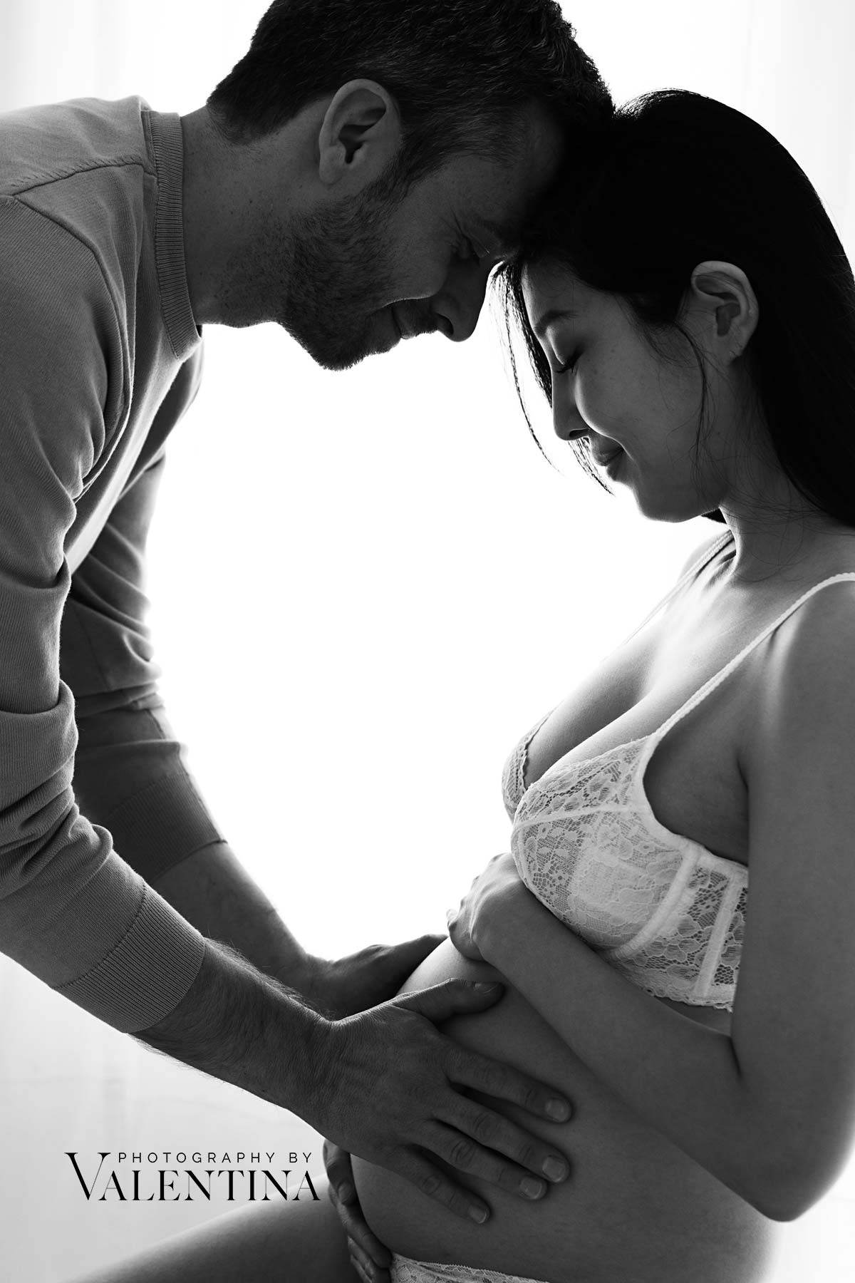Black and white portrait of a pregnant woman and her partner, both gently holding her baby bump with their foreheads touching, capturing an intimate and emotional moment of connection during maternity