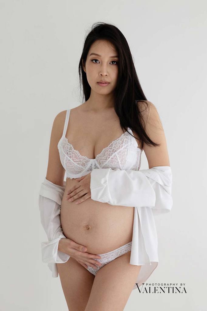 Pregnant woman in her third trimester holding her bump while wearing a white shirt – calm and intimate studio portrait.