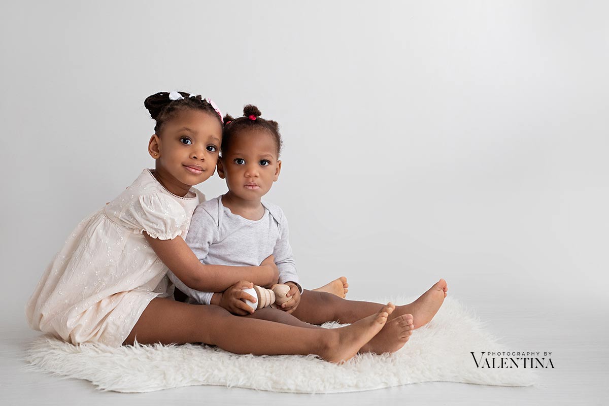 Two young sisters sitting on a white backdrop, holding a baby doll together.