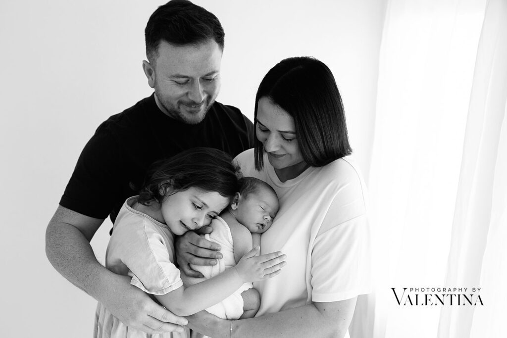 Family portrait: mum is holding her newborn on her chest while big sister is leaning on her baby sister and dad is looking at them with pride.