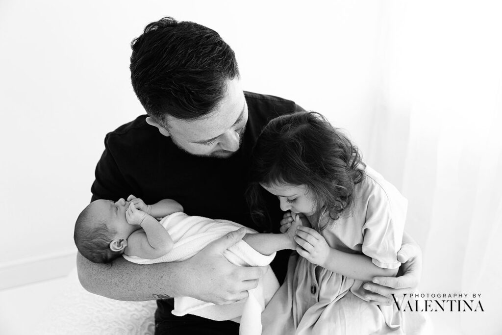 Dad and his two girls. Big sister kidding newborn foot. Sweet moment of storytelling.