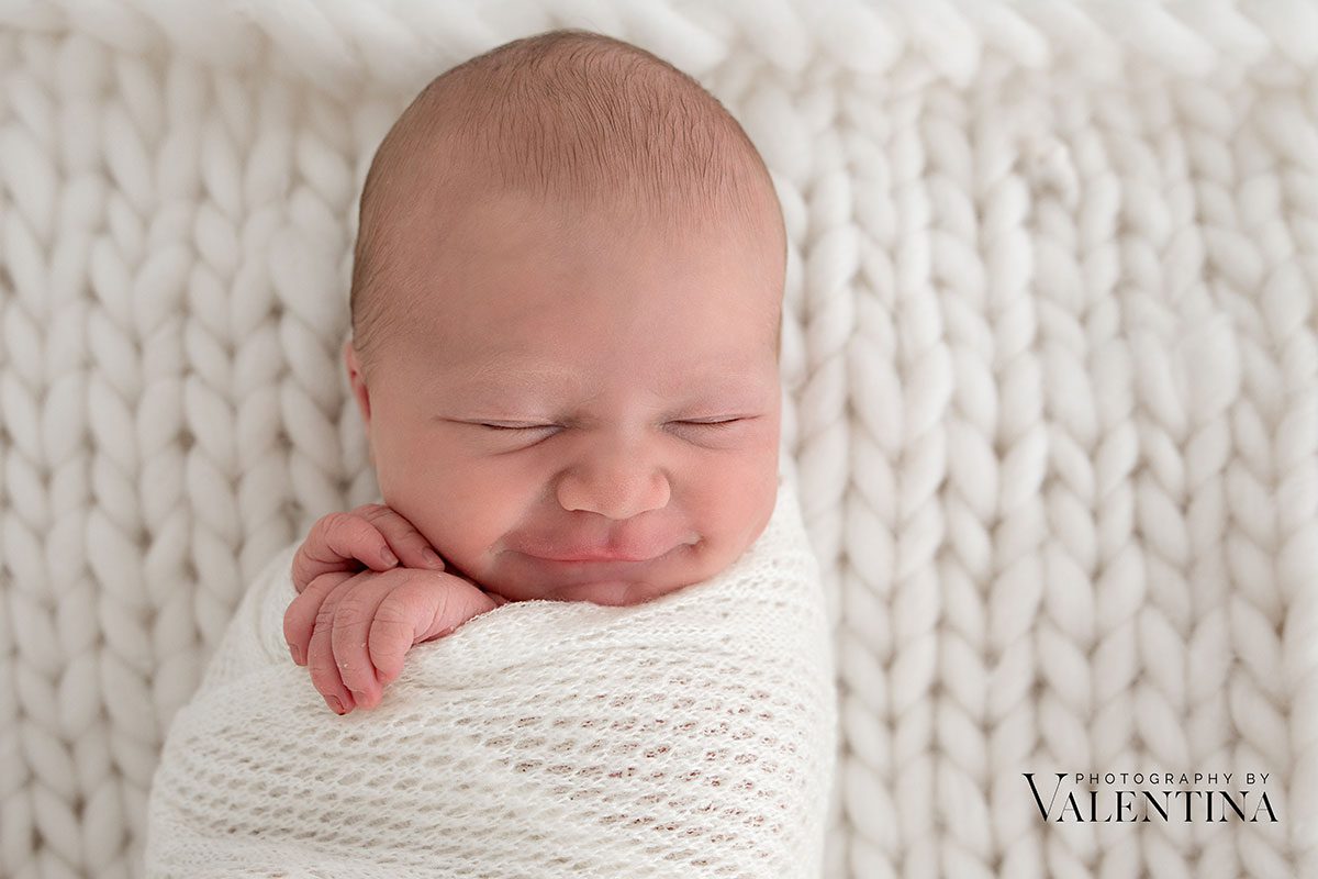 Close-up of newborn baby wrapped in a white knit, gently frowning in sleep on a chunky knitted background